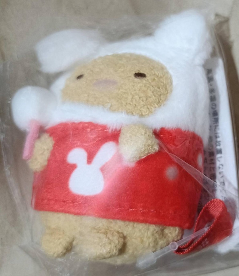 とんかつキャラのてのりぬいぐるみ正面。きつね色もこもこボディに白フード付きコート、赤衣装にうさぎマークプリント、ピンクのキャンディスティックを持つ冬スタイル。